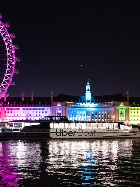Uber Boat on Thames River at night with London Eye and illuminated buildings in background.