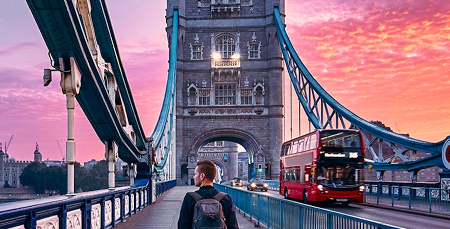 Person walking on Tower Bridge in London at sunset with a red double-decker bus passing by.