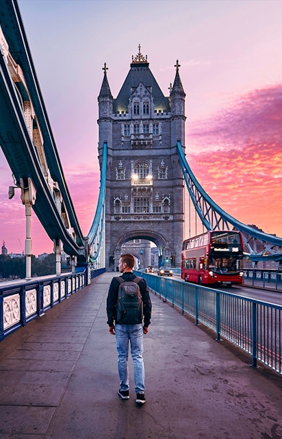 Person walking on Tower Bridge in London at sunset with a red double-decker bus passing by.