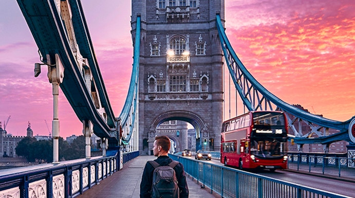 Person walking on Tower Bridge in London at sunset with a red double-decker bus passing by.