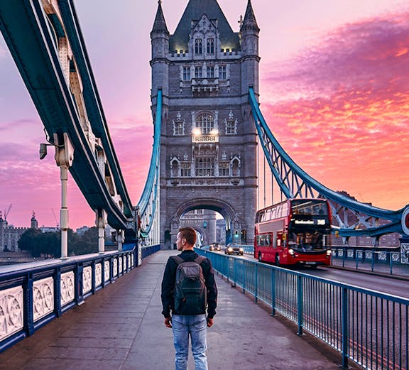 Person walking on Tower Bridge in London at sunset with a red double-decker bus passing by.