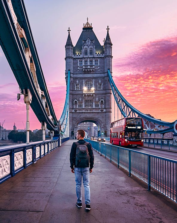 Person walking on Tower Bridge in London at sunset with a red double-decker bus passing by.