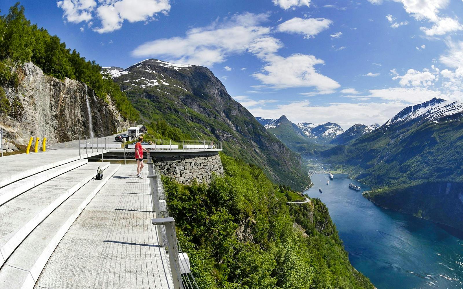Viewing platform overlooking Geirangerfjord with cruise ships, mountains, and a waterfall in Ålesund, Norway.