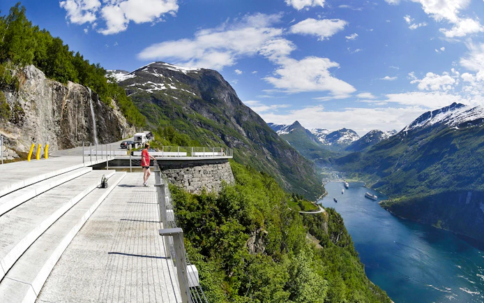 Viewing platform overlooking Geirangerfjord with cruise ships, mountains, and a waterfall in Ålesund, Norway.