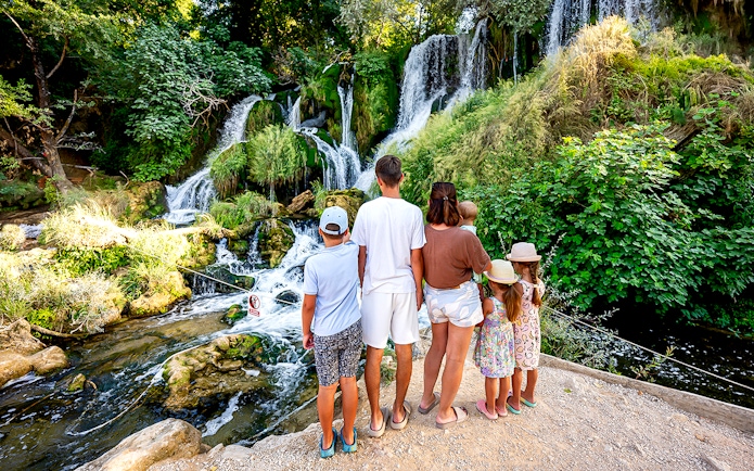 Family observing Kravica Waterfalls surrounded by lush greenery.