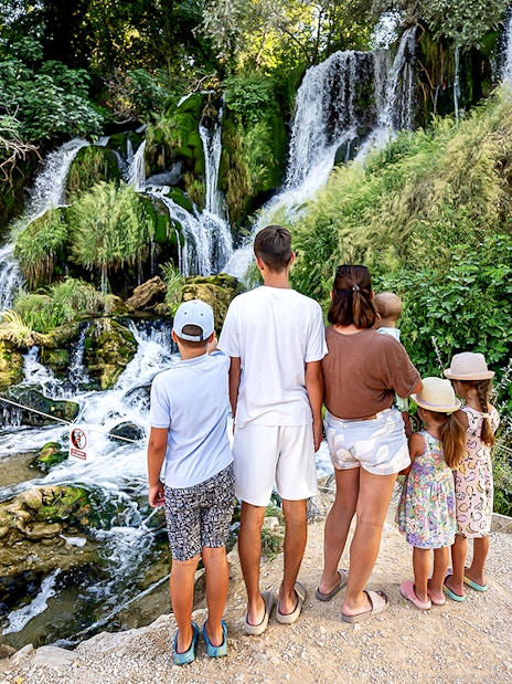 Family observing Kravica Waterfalls surrounded by lush greenery.