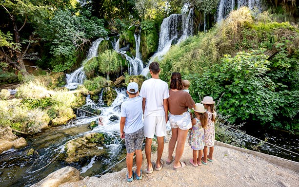 Family observing Kravica Waterfalls surrounded by lush greenery.
