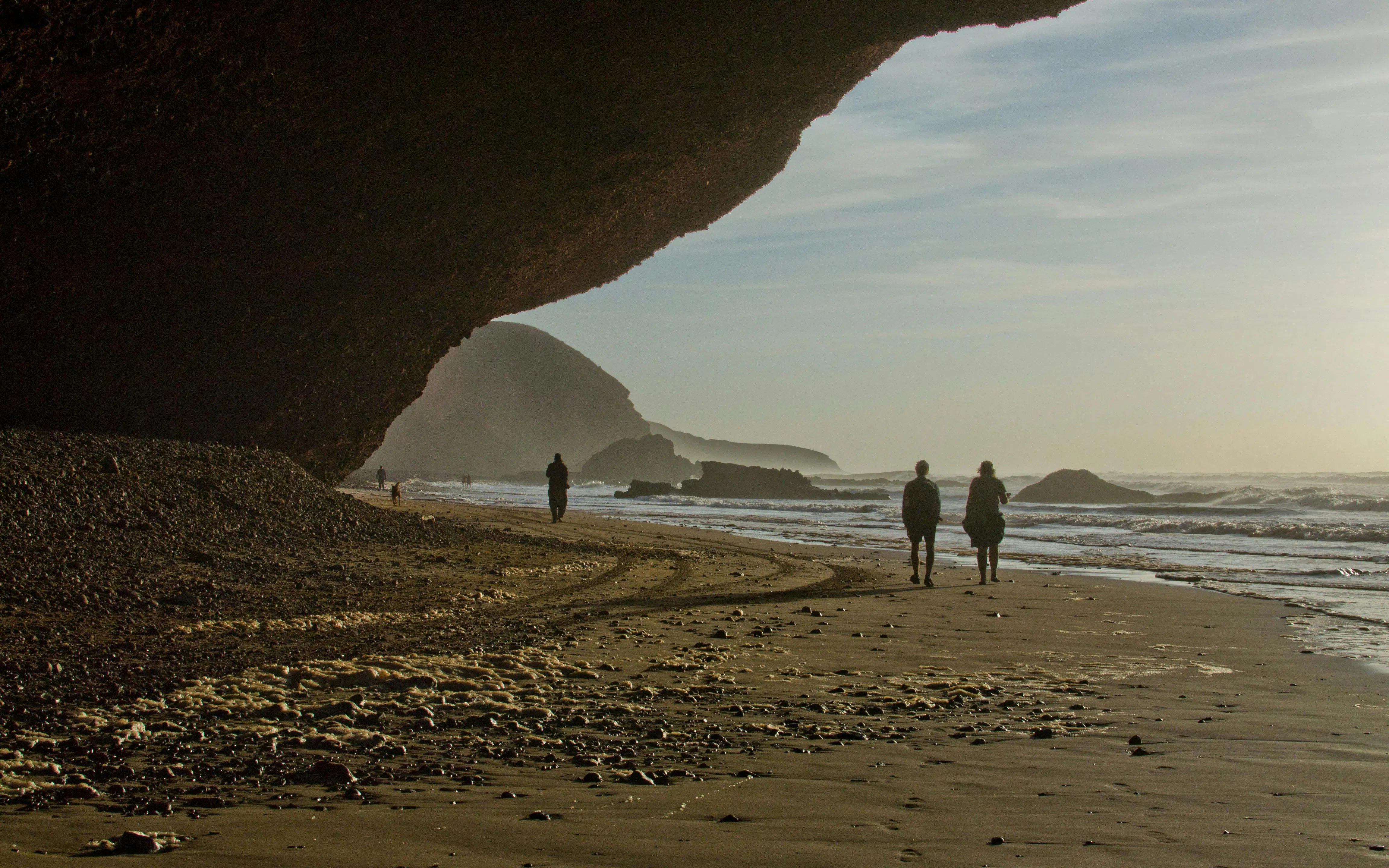 People walking along Legzira Beach at sunset, Morocco, with natural rock arch and ocean waves.