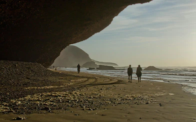 People walking along Legzira Beach at sunset, Morocco, with natural rock arch and ocean waves.