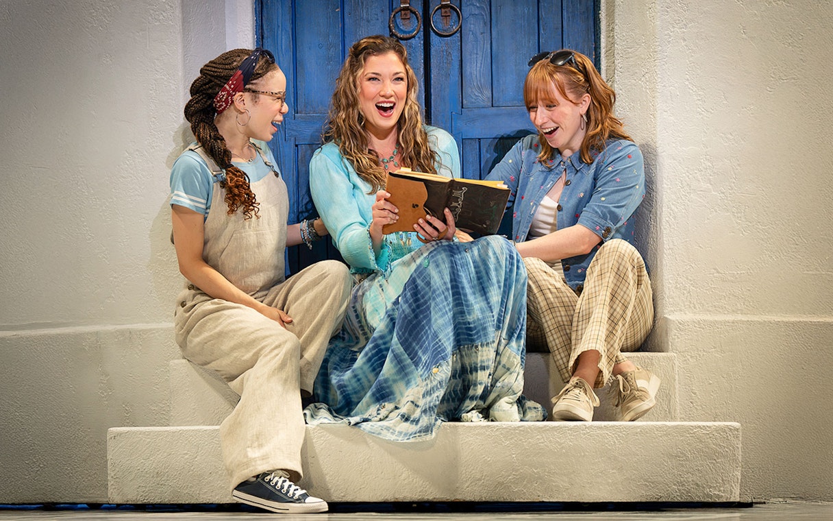 Three women sitting on steps, joyfully reading a book, related to Mamma Mia! musical.