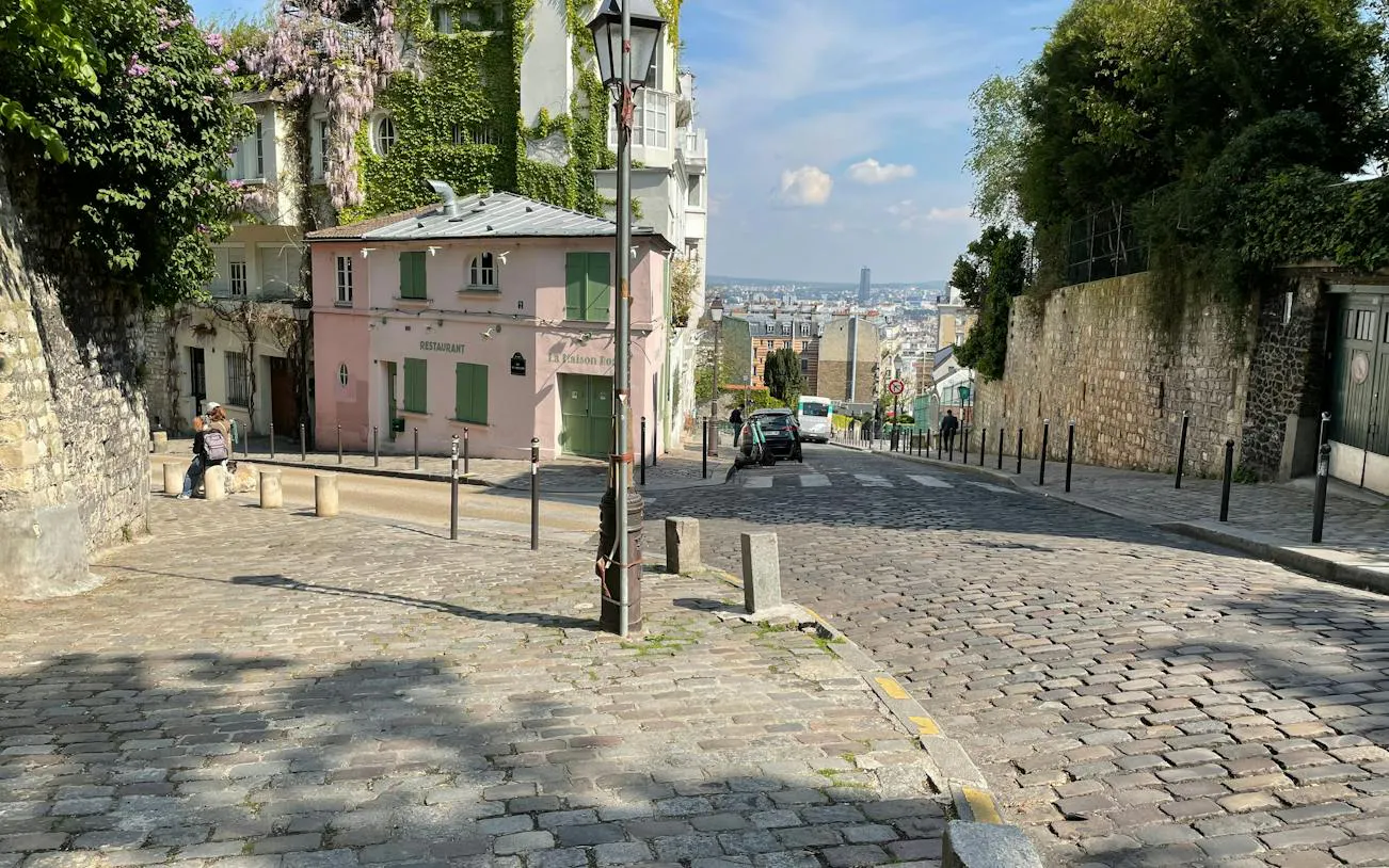 Cobblestone street in Montmartre, Paris, with ivy-covered buildings and distant city view.