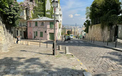 Cobblestone street in Montmartre, Paris, with ivy-covered buildings and distant city view.