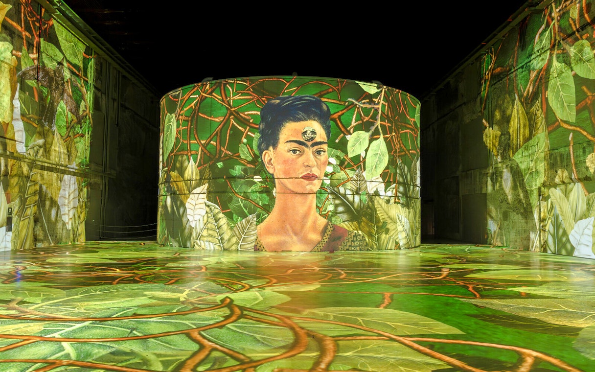 Frida Kahlo artwork projected at Bassins Des Lumieres exhibition, surrounded by vibrant foliage.