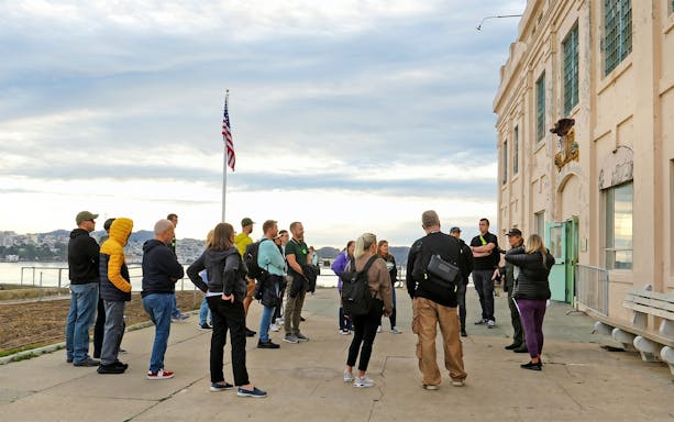 Group on guided tour outside Alcatraz Island building, San Francisco Bay in background.