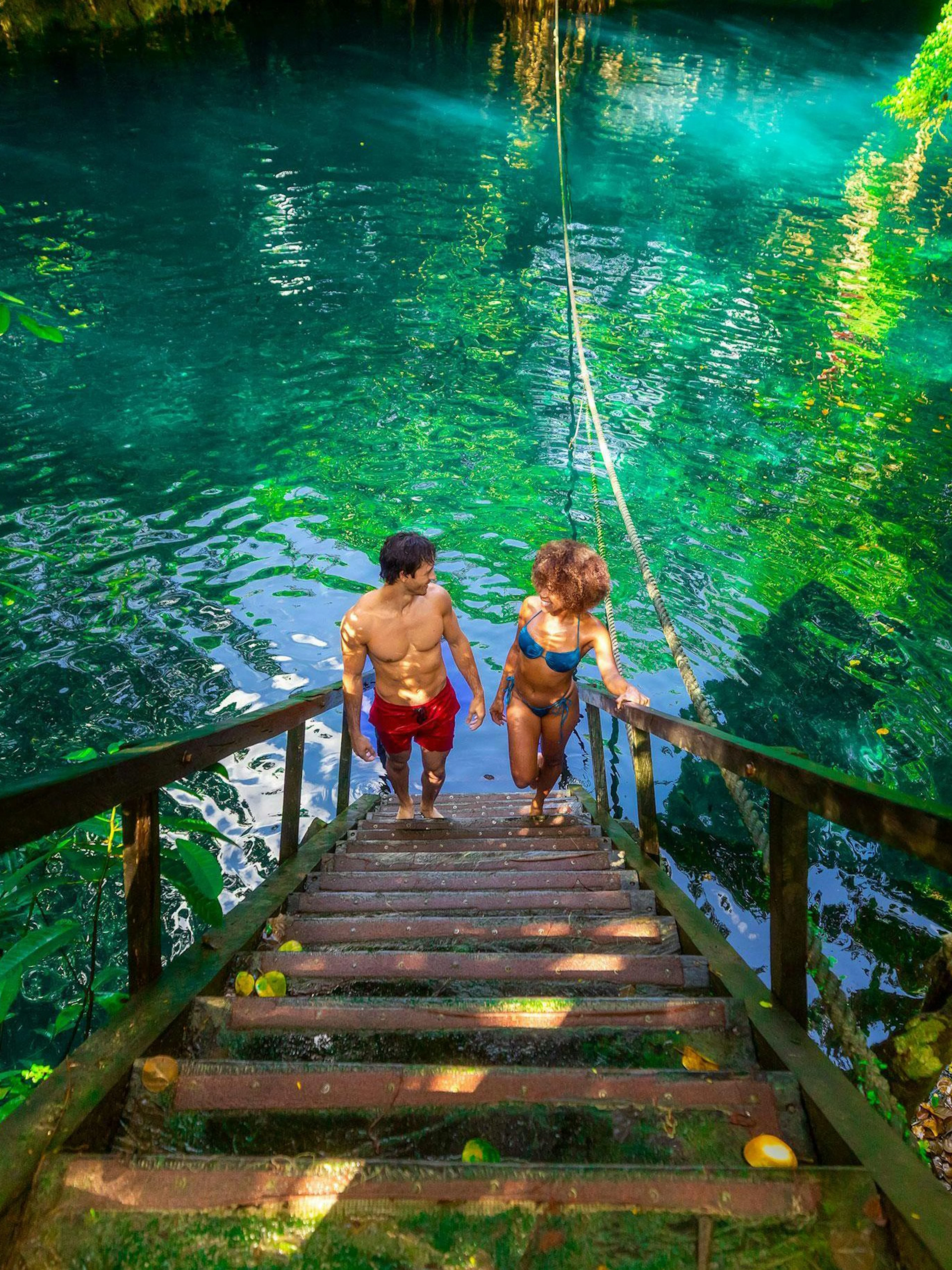 Visitors descending stairs into a cenote in Cancun, surrounded by lush greenery and clear water.