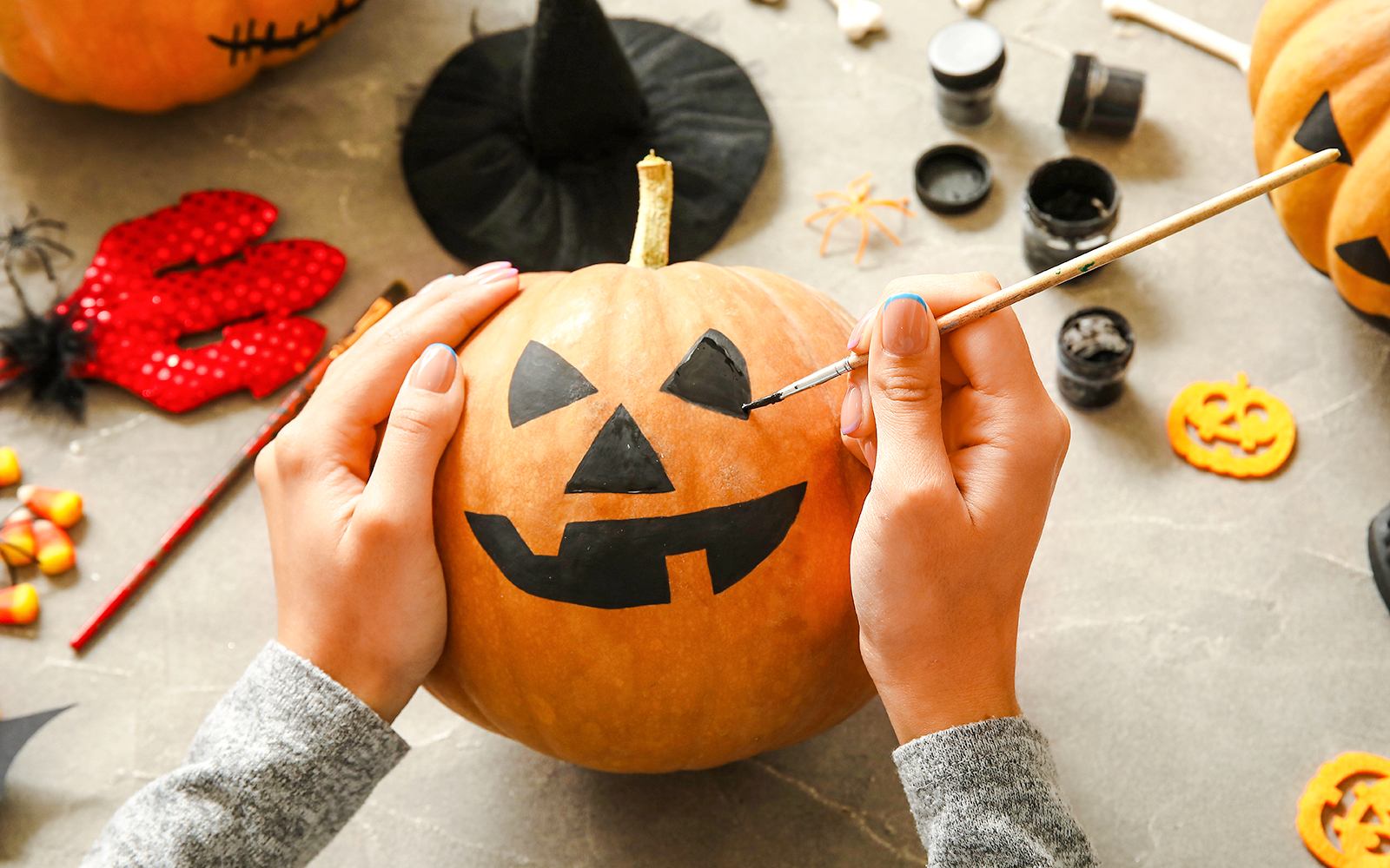 Woman painting jack-o'-lantern face on pumpkin at grey table.