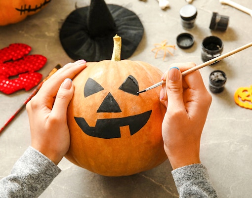 Woman painting jack-o'-lantern face on pumpkin at grey table.