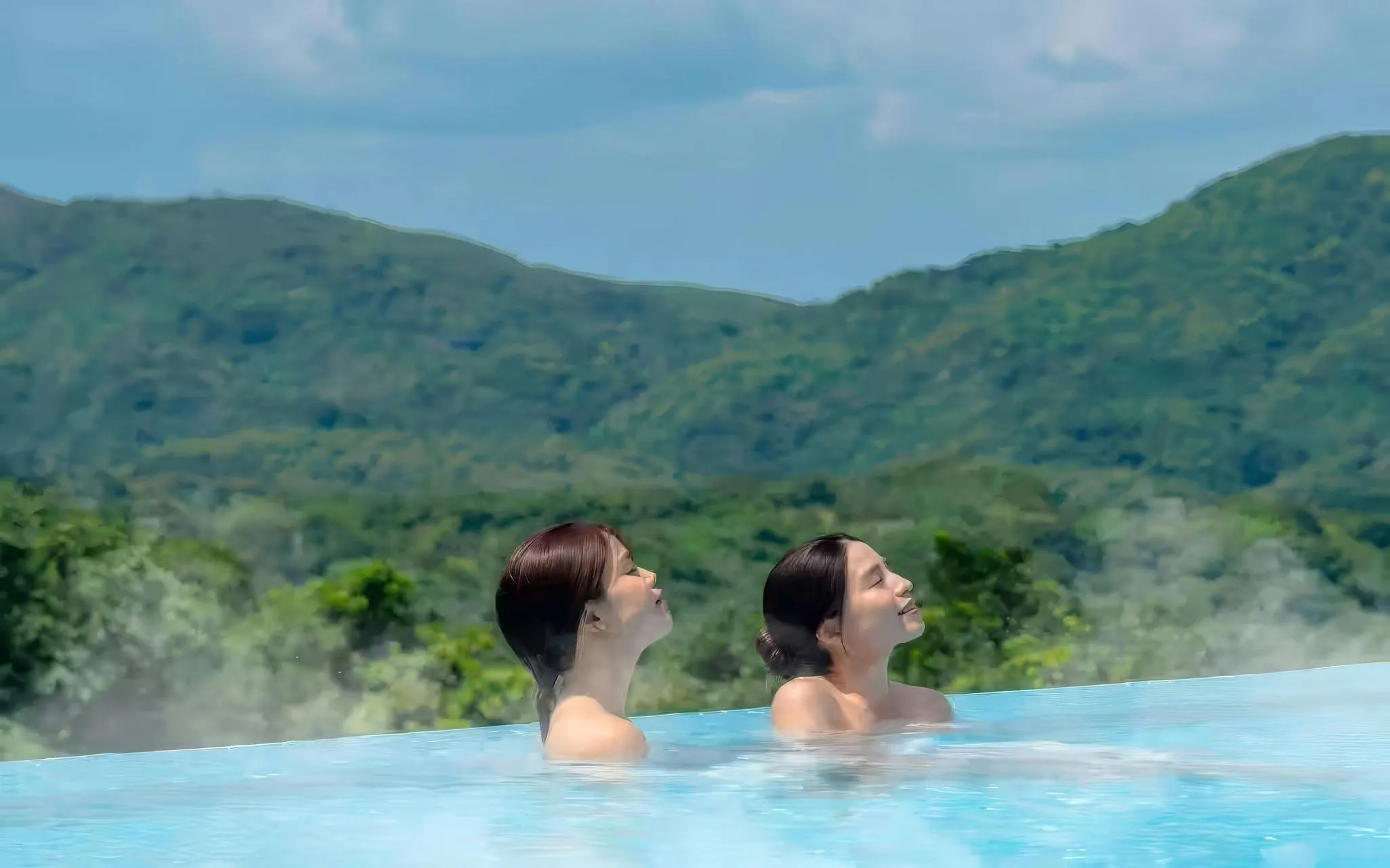 Two women relaxing in an infinity pool at SPA JUNGLIA Hot Spring with mountain views.