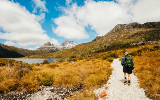 Hiker on trail in Cradle Mountain National Park, Tasmania, with rugged peaks and lake in view.