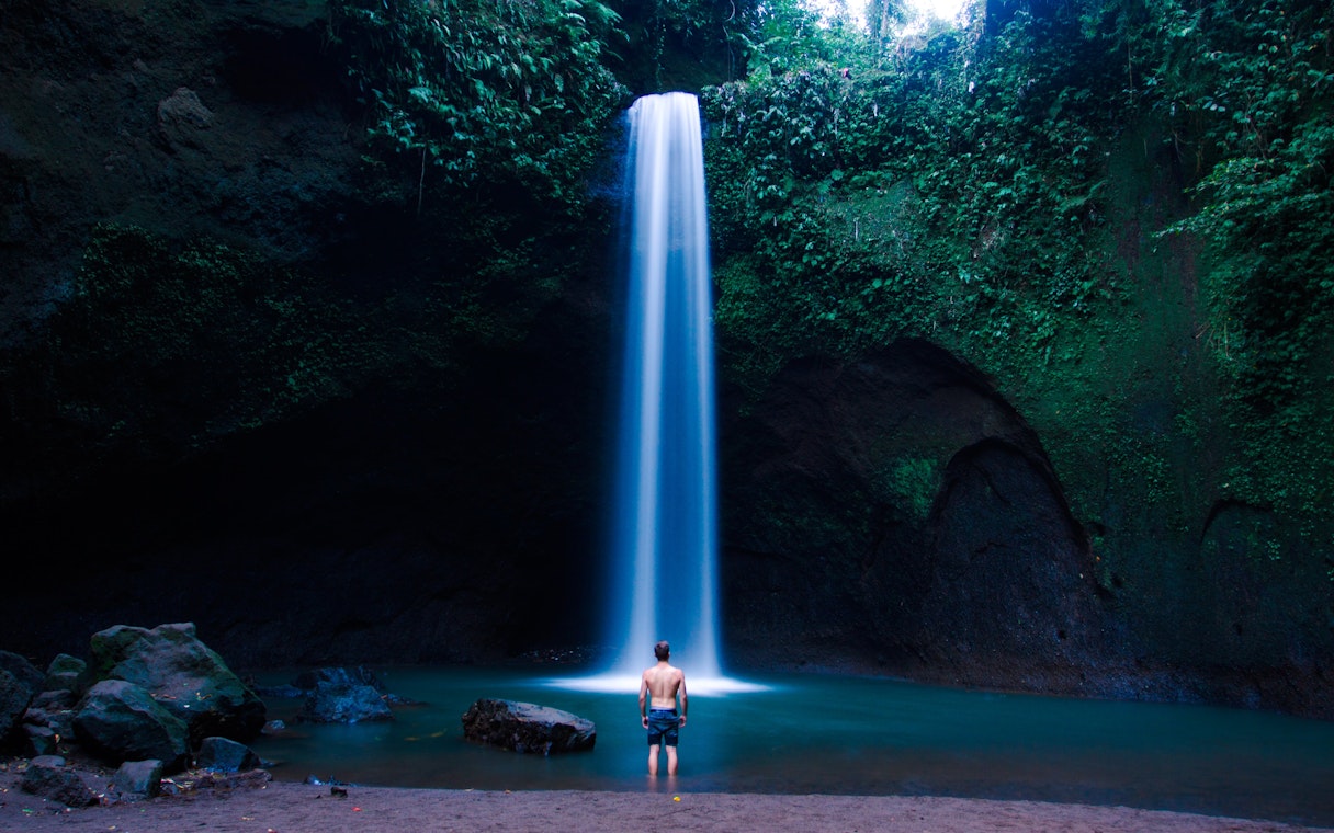 Tourist standing at the base of Tibumana Waterfall in Bali, surrounded by lush greenery.