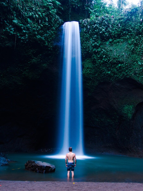 Tourist standing at the base of Tibumana Waterfall in Bali, surrounded by lush greenery.