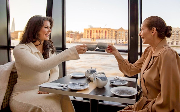 Two people enjoying tea on a Thames River cruise with cityscape view.