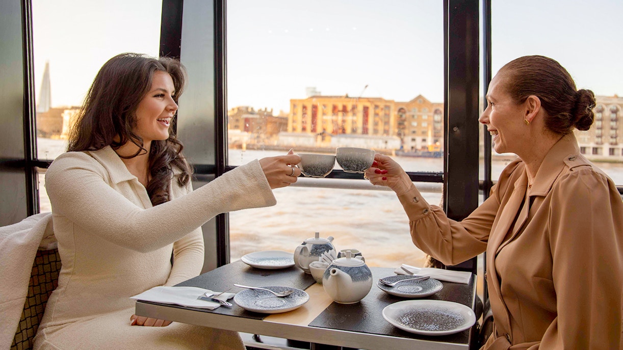 Two people enjoying tea on a Thames River cruise with cityscape view.