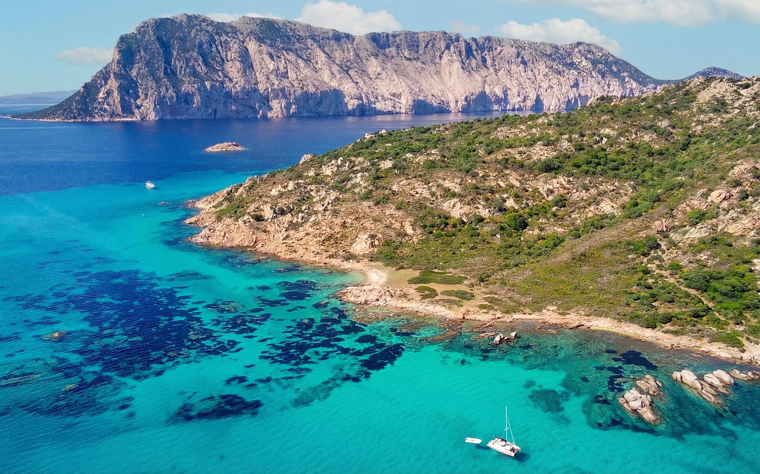 Aerial view of Tavolara Island's rocky coastline and turquoise waters with a sailboat.
