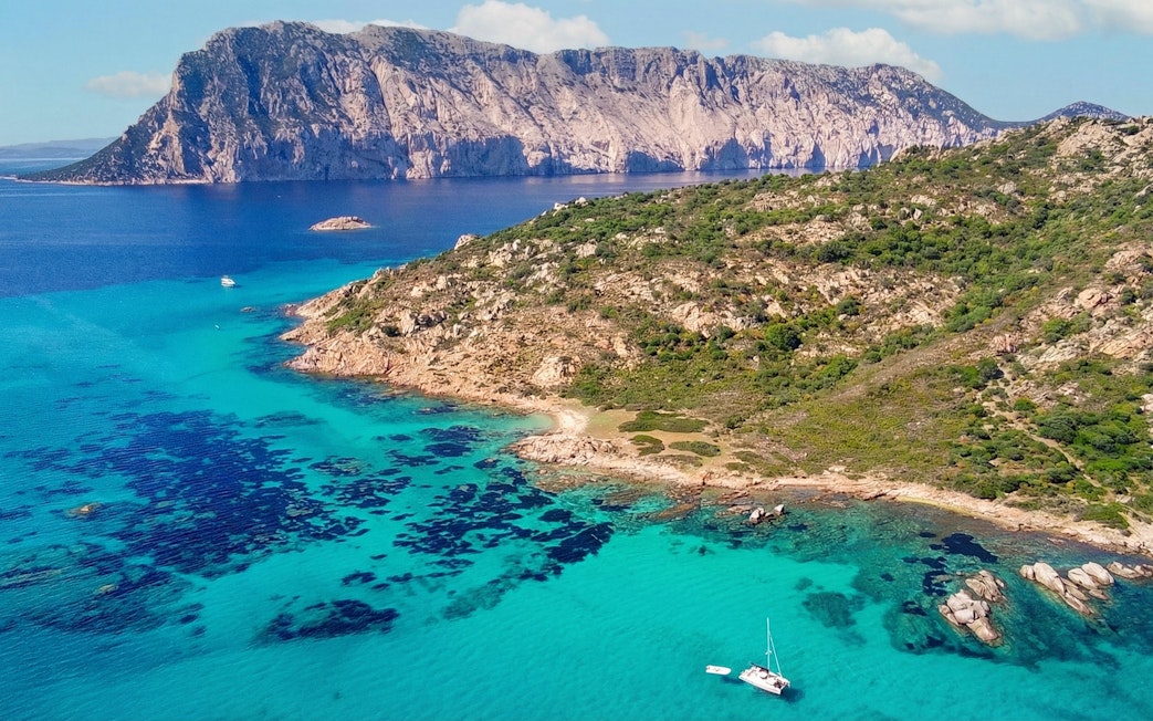 Aerial view of Tavolara Island's rocky coastline and turquoise waters with a sailboat.