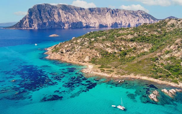 Aerial view of Tavolara Island's rocky coastline and turquoise waters with a sailboat.