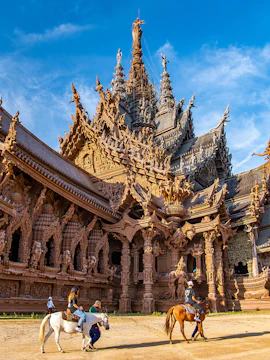 Visitors on horseback in front of the intricate wooden Sanctuary of Truth in Pattaya, Thailand.