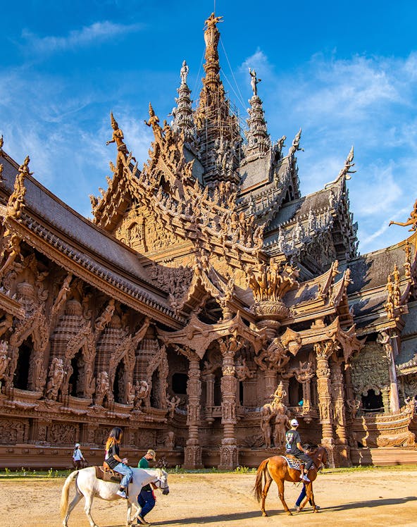 Visitors on horseback in front of the intricate wooden Sanctuary of Truth in Pattaya, Thailand.