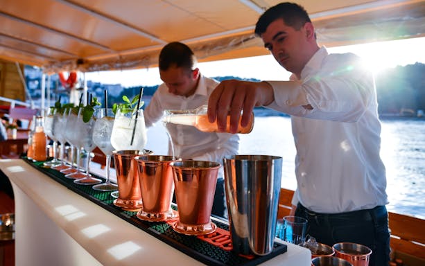 Mixologist pouring cocktail into copper mugs on a boat with a scenic view.