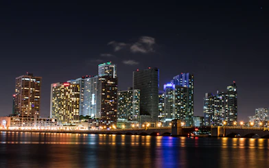 Miami skyline at night with illuminated skyscrapers and bridge reflecting on the water.