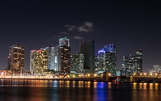 Miami skyline at night with illuminated skyscrapers and bridge reflecting on the water.