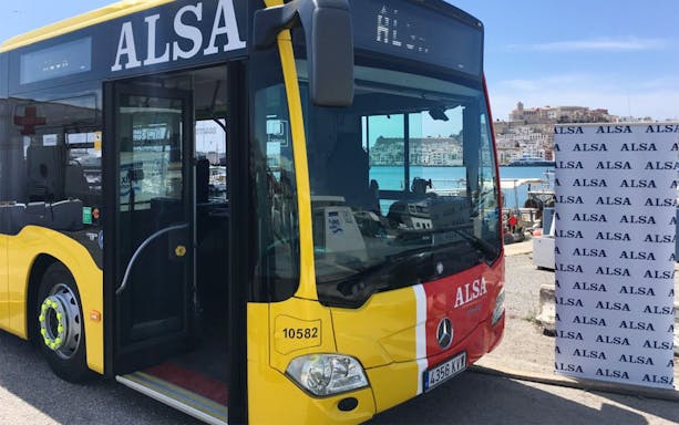 Yellow ALSA bus at Ibiza port with city view in background.