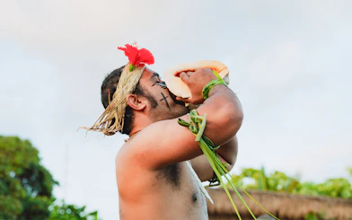 Man blowing conch shell at Mauka Warriors Luau, wearing traditional attire with face paint.