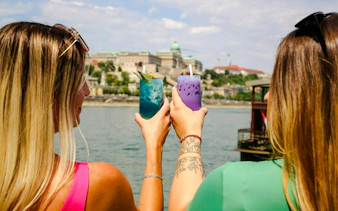 Guests holding colorful drinks on Danube Sightseeing Cruise with Budapest in background.