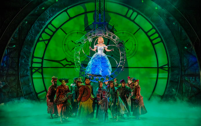 Stage scene from the musical Wicked with performers in front of a large clock.