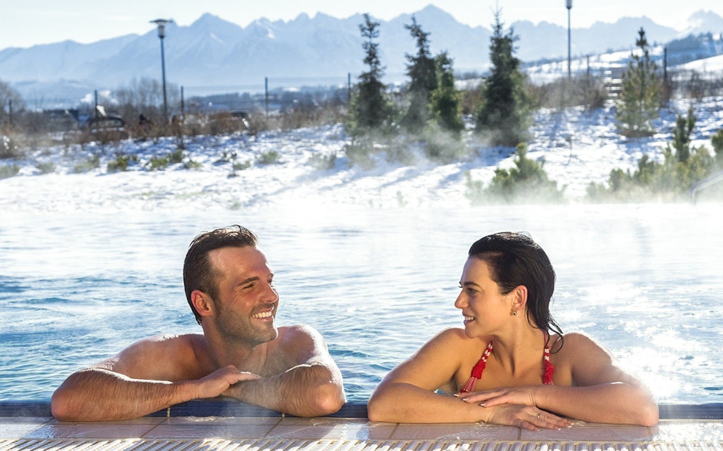 Tourists relaxing in a thermal spa with snowy Zakopane mountains in the background.
