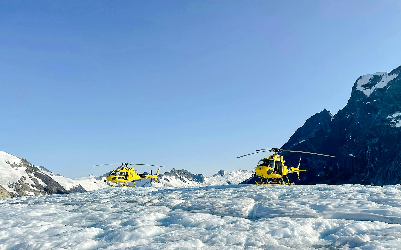 Yellow helicopters on a glacier during Juneau helicopter tour with mountain backdrop.