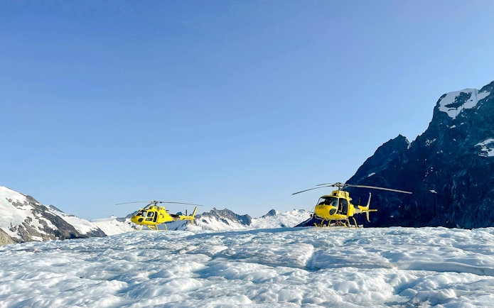 Yellow helicopters on a glacier during Juneau helicopter tour with mountain backdrop.