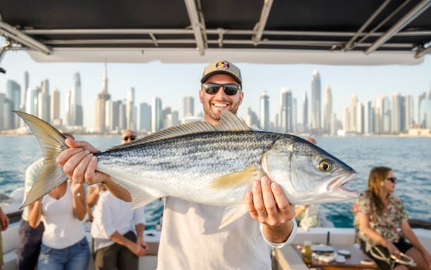 Man holding a large fish on a sports fishing cruise with Dubai skyline in the background.