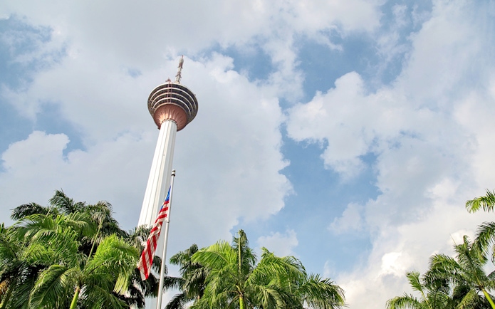 KL Tower in Kuala Lumpur with palm trees and Malaysian flag in foreground.