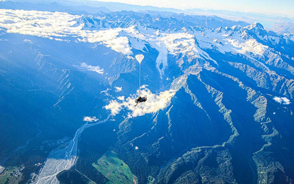 Skydiver over Franz Josef Glacier, New Zealand, with snow-capped mountains and lush valleys below.