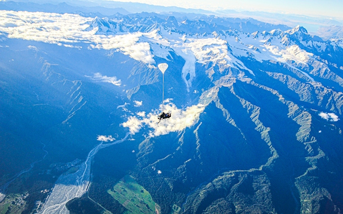Skydiver over Franz Josef Glacier, New Zealand, with snow-capped mountains and lush valleys below.