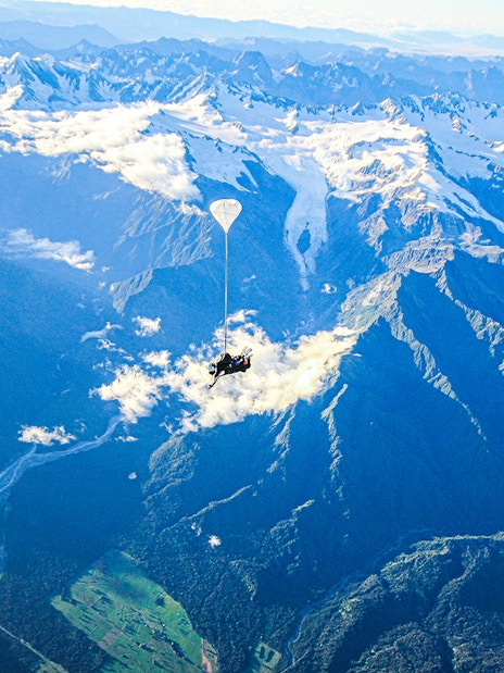 Skydiver over Franz Josef Glacier, New Zealand, with snow-capped mountains and lush valleys below.