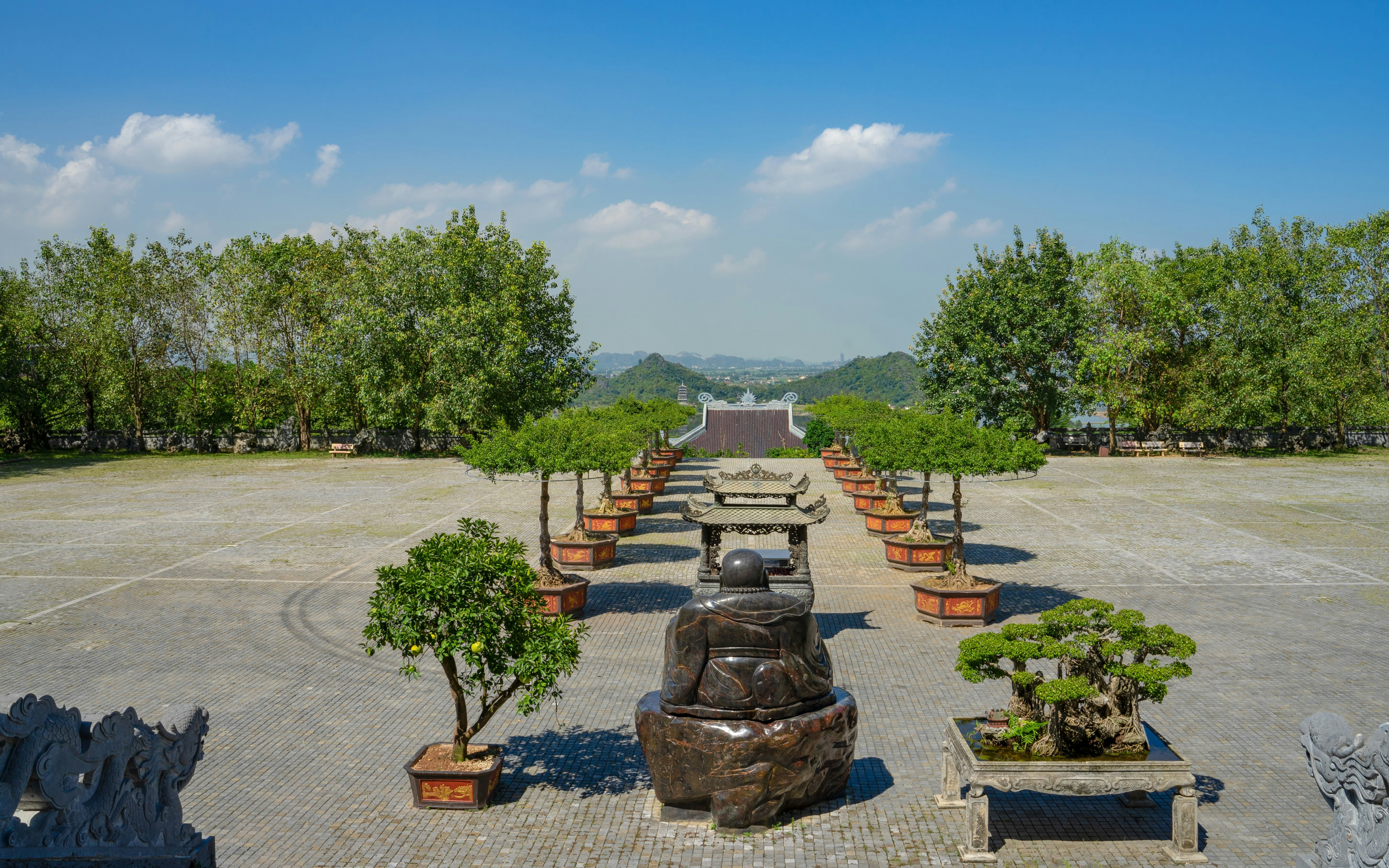 Buddhist statue and bonsai trees at Bai Dinh Pagoda complex under blue sky, Vietnam.