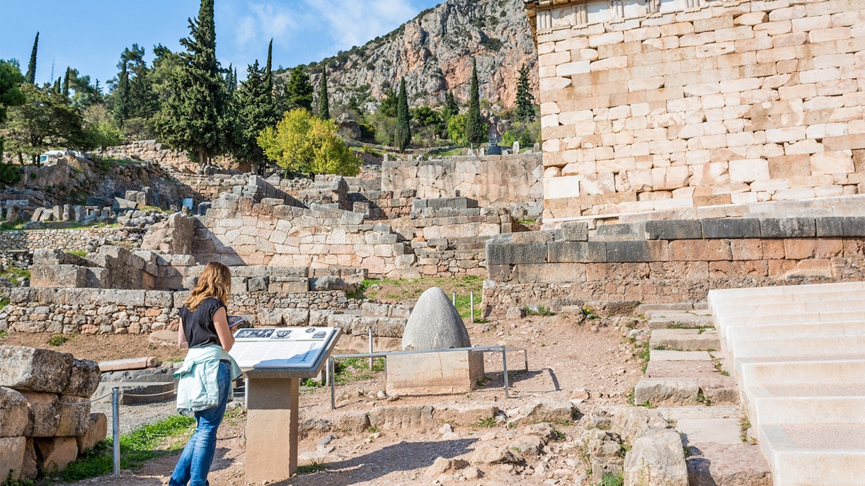 Visitor reading information at Delphi archaeological site, Greece, with ancient ruins and mountains in the background.