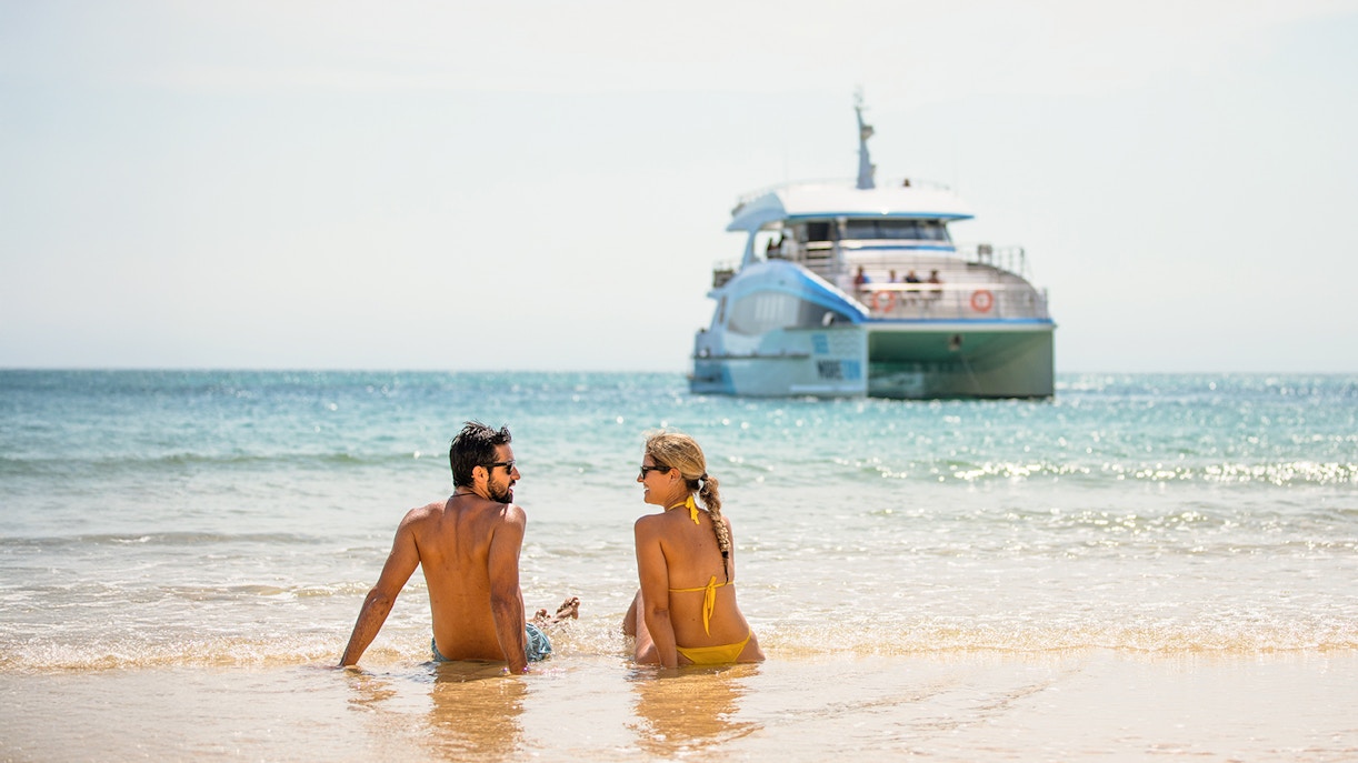 Couple sitting on Moreton Island beach with a boat in the background.